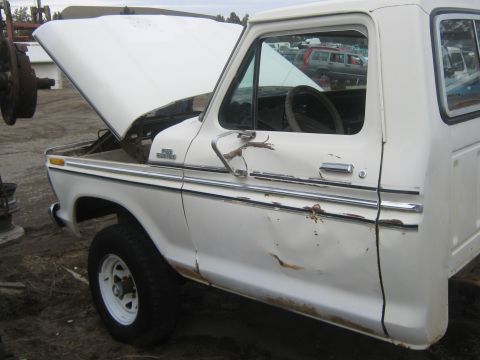 1978 Ford Bronco - A white pickup truck sits in a junkyard with its hood open, showing signs of damage and wear.