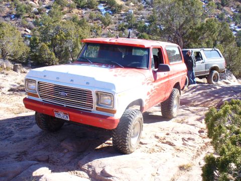 1979 Ford Bronco - A red and white 1979 Ford Bronco with flame decals navigates rocky terrain, with another SUV visible in the background.