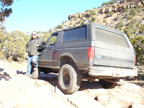 1979 Ford Bronco - A man stands next to a dark gray SUV on a dusty, rocky trail under a clear blue sky.