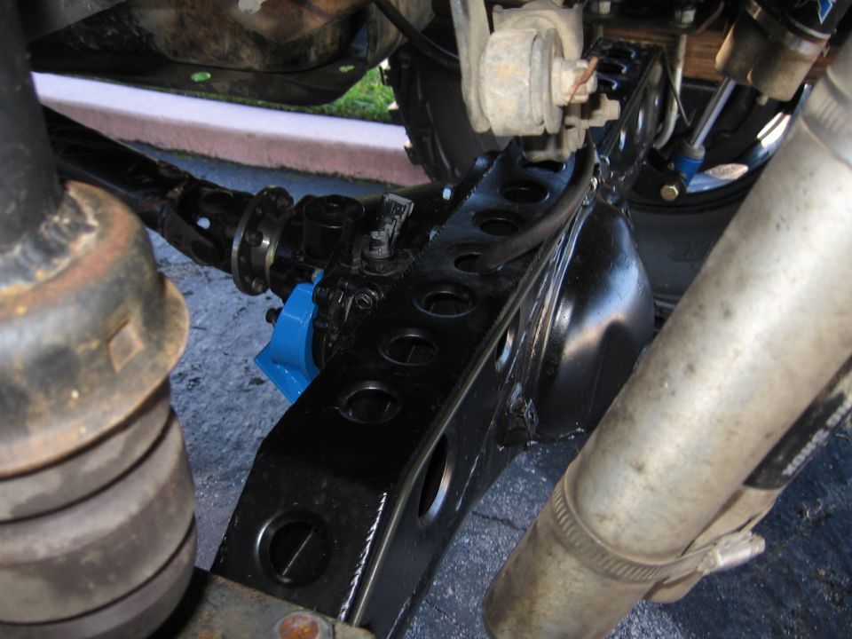 Close-up of a black rear axle housing with a blue bracket and a silver exhaust pipe. Supermotors.net logo visible