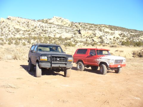 1979 Ford Bronco - Two Ford Broncos, one red with flame decals and a dark-colored one, are parked in a dry, rocky terrain under a clear blue sky.