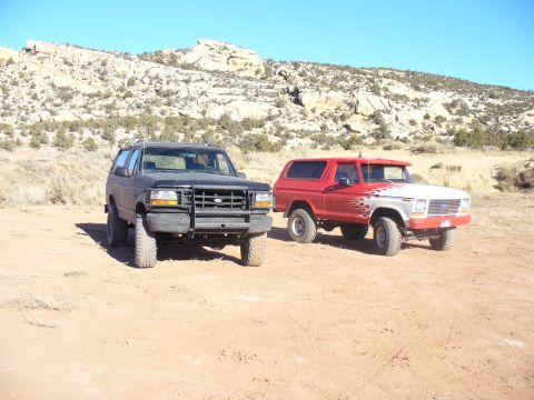 1979 Ford Bronco - Two SUVs, one red and white with flame decals and another black, sit parked in a dry, rocky landscape under a clear blue sky.