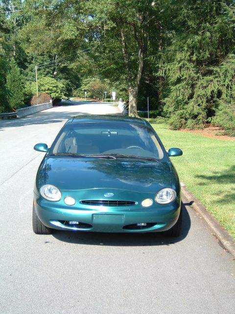 1997 Ford Taurus - A green 1997 Ford Taurus is parked on the side of a road with trees in the background.