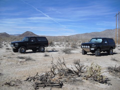 1986 Ford Bronco - Two black Ford Broncos are parked in a dry, desert environment with mountains in the background.