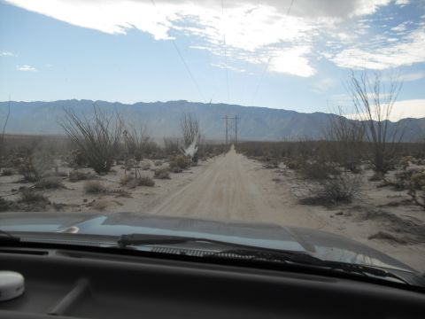 1986 Ford Bronco - View from inside a vehicle on a dirt road in a desert with mountains and power lines under a cloudy sky.