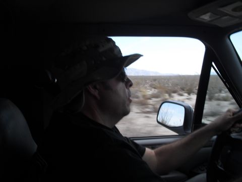 1986 Ford Bronco - A man wearing a camouflage hat drives a vehicle through a barren desert, with mountains in the distance.