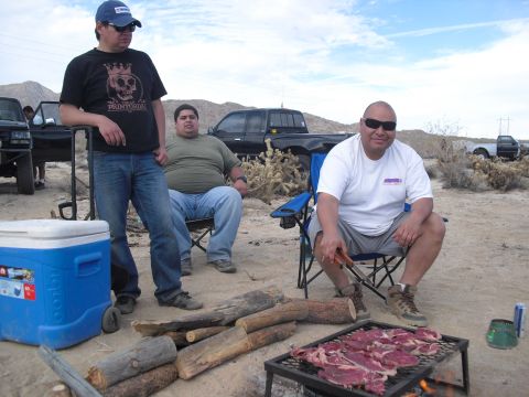 1986 Ford Bronco - Men grill meat at a desert campsite with trucks in the background.