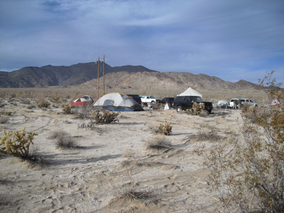 Desert campsite with tents and several vehicles parked around them. Mountains in the background