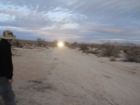 1986 Ford Bronco - A person stands on a dirt road in the desert, looking towards distant headlights as the sky darkens.