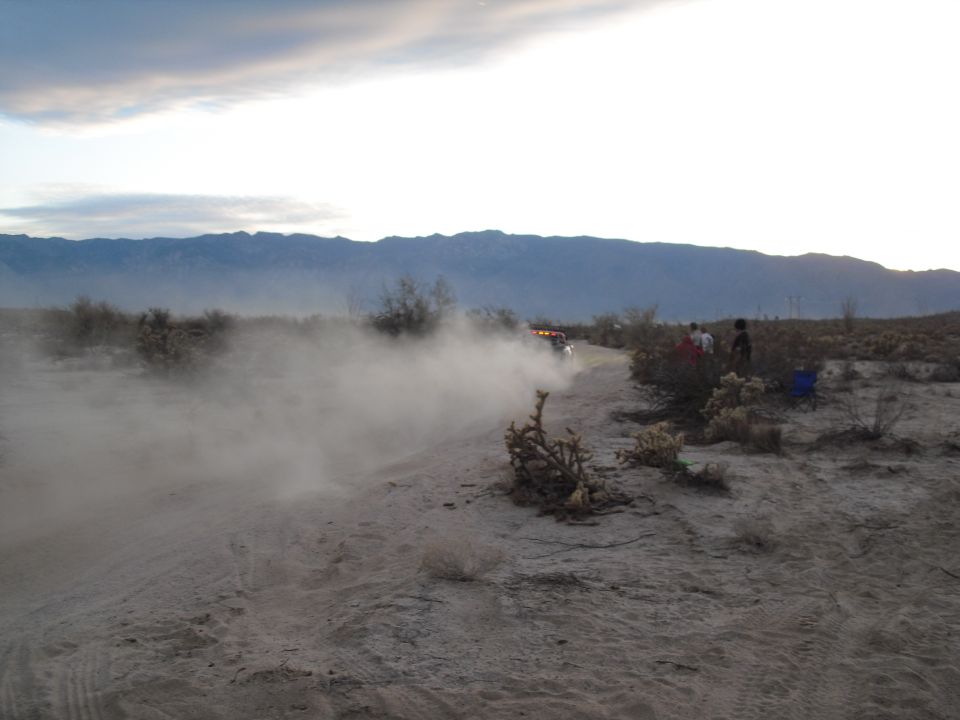 Off-road vehicle driving on a dirt track, kicking up a large dust cloud. Mountains in the background