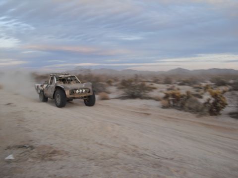 1986 Ford Bronco - A custom off-road truck navigates a dusty desert course under a cloudy sky.
