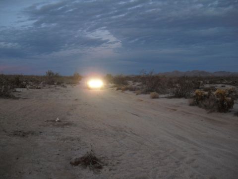 1986 Ford Bronco - A vehicle drives down a dusty desert road as the sun sets, its headlights cutting through the twilight.