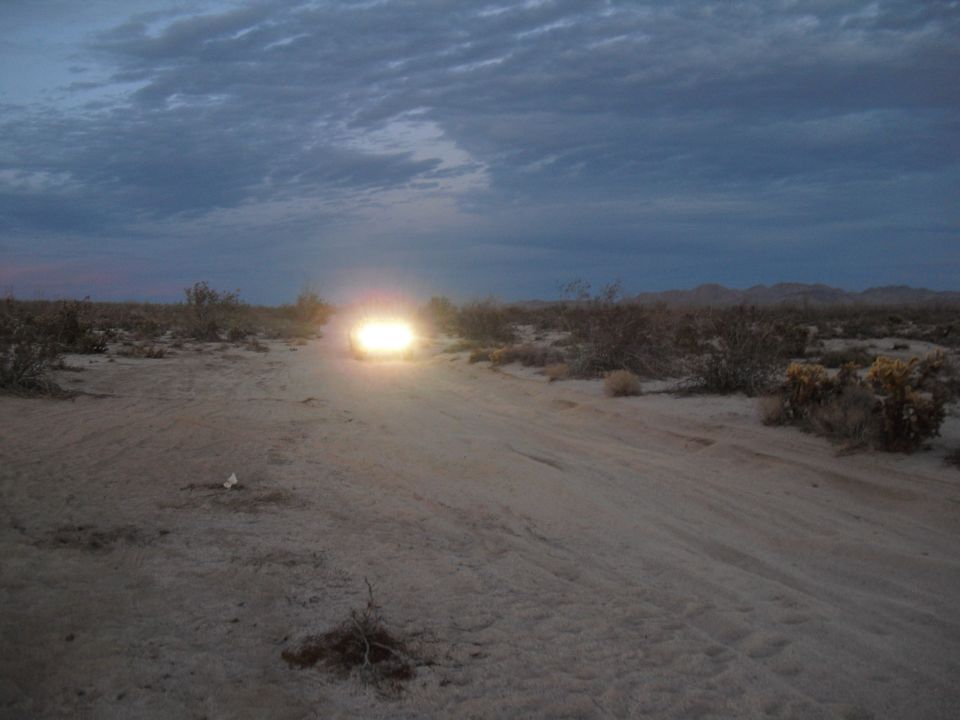 Vehicle driving on dirt road in desert at dusk, headlights on