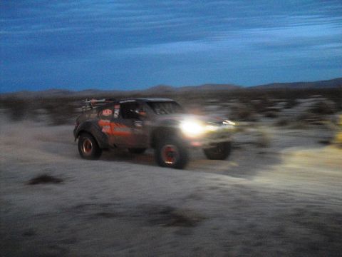 1986 Ford Bronco - A modified off-road truck races across a desert landscape as the sun sets.