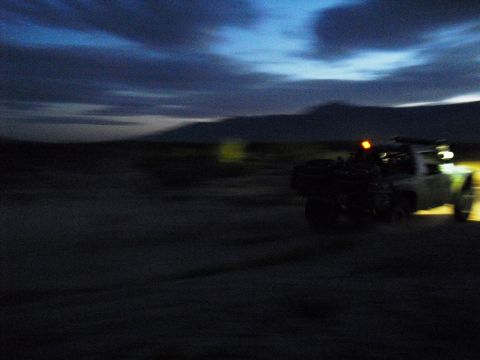 1986 Ford Bronco - A vehicle drives through a desert landscape as the sun sets, its lights illuminating the path ahead.