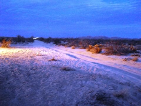 1986 Ford Bronco - Headlights cut through the twilight of a desert landscape, illuminating tire tracks on a dirt road.