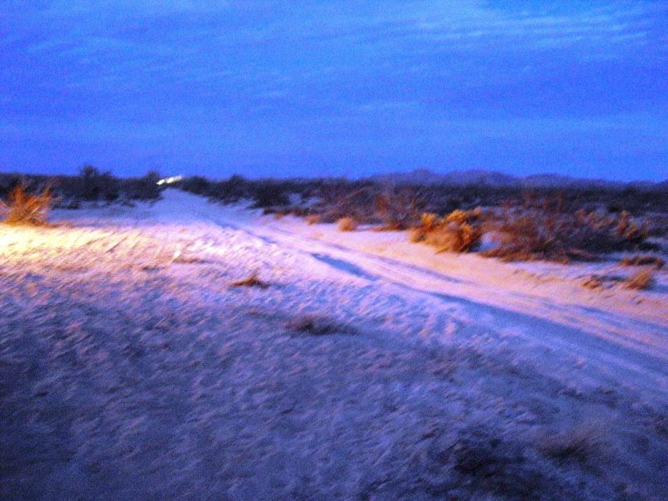 Desert landscape at dusk with tire tracks and distant headlights illuminating the sandy terrain