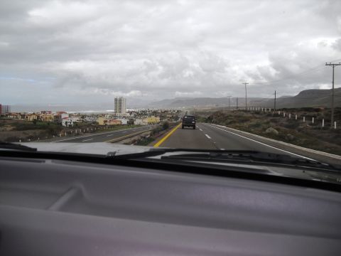 1986 Ford Bronco - A black SUV travels down a highway towards a coastal town under a cloudy sky.