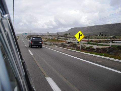 1986 Ford Bronco - A black SUV travels down a highway under a cloudy sky, with a yellow directional sign visible on the right.