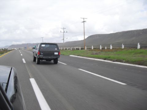 1986 Ford Bronco - A black SUV travels down a highway under an overcast sky, with another vehicle in the foreground.