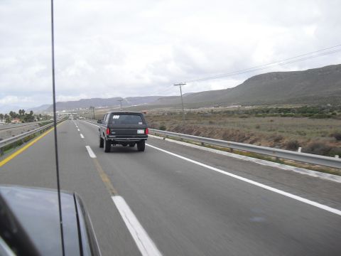 1986 Ford Bronco - A black SUV travels down a highway under a cloudy sky, with a mountainous landscape visible in the distance.