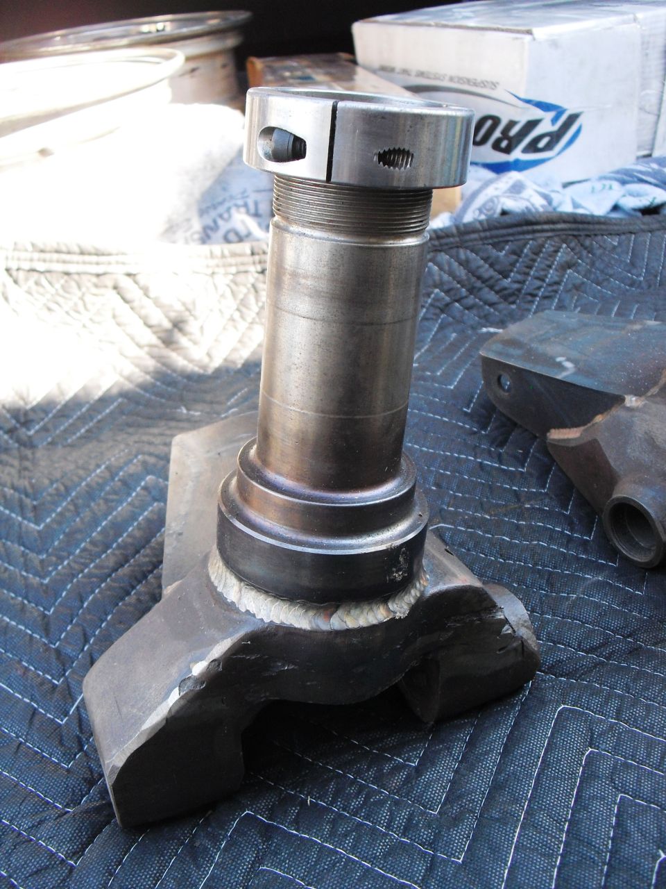 Close-up of a metal spindle and knuckle assembly, possibly for a truck or SUV, resting on a quilted mat. A box with 'PRO' visible is in the background
