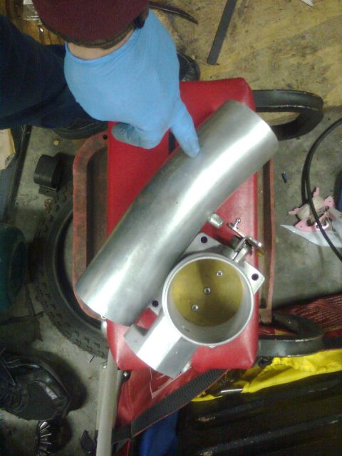 1984 Ford Bronco - A gloved hand points to an aluminum intake pipe positioned above a throttle body, resting on a red container.