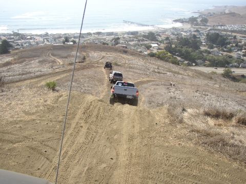1989 Ford Bronco - Multiple vehicles navigate a dusty trail overlooking a coastal town and the ocean.