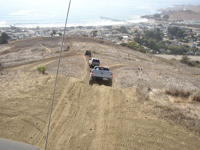 Silver pickup and other vehicles descending a sandy off-road hill toward a coastal town and ocean