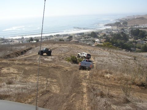 1989 Ford Bronco - Multiple off-road vehicles traverse a dry, grassy hillside with the ocean and a coastal town in the background.