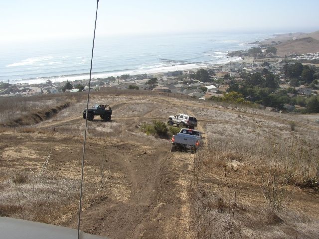 Three off-road vehicles on a dirt hilltop trail above a coastal town and ocean