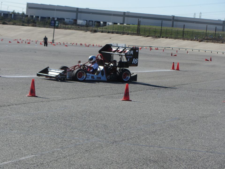 Racing go-kart with driver wearing helmet, number 16 on rear wing, on asphalt track with orange cones