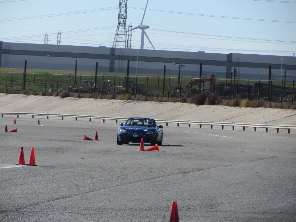 Blue convertible sports car navigating a cone course on a track. Power lines and industrial buildings in the background