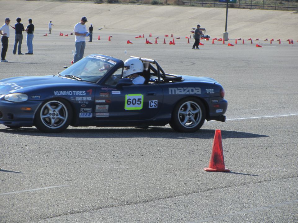 Blue Mazda Miata with racing decals and number 605 on the side, seen from the side at a track event