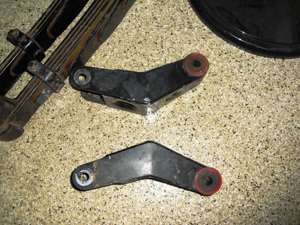 Leaf spring assembly and two black metal shackle parts with red bushings on speckled floor