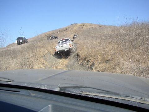 1989 Ford Bronco - Several vehicles navigate a challenging off-road course on a sunny day, with one pickup truck descending a steep, rutted path.