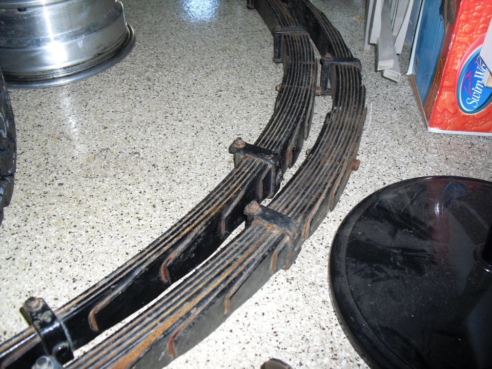 Pair of old leaf springs with rust, sitting on a speckled floor next to a tire rim and a black round object