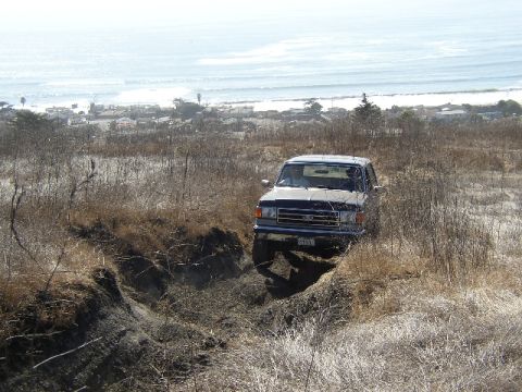 1989 Ford Bronco - A dark blue Ford Bronco navigates an off-road trail with the ocean in the background.