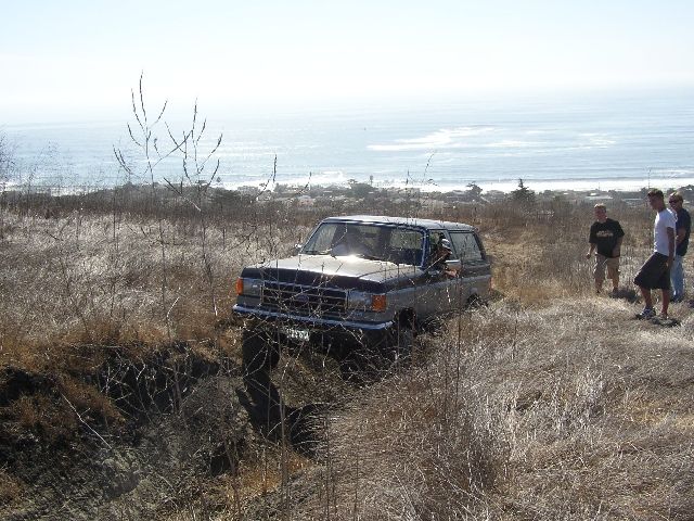 Maroon Ford Bronco stuck nose-down in a gully on a coastal hillside with three onlookers.