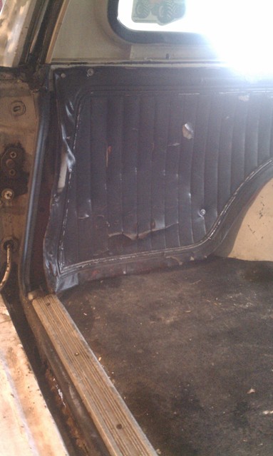 1978 Ford Bronco - The interior of a vehicle displays damaged black paneling and a dirty floor with a wooden trim piece.