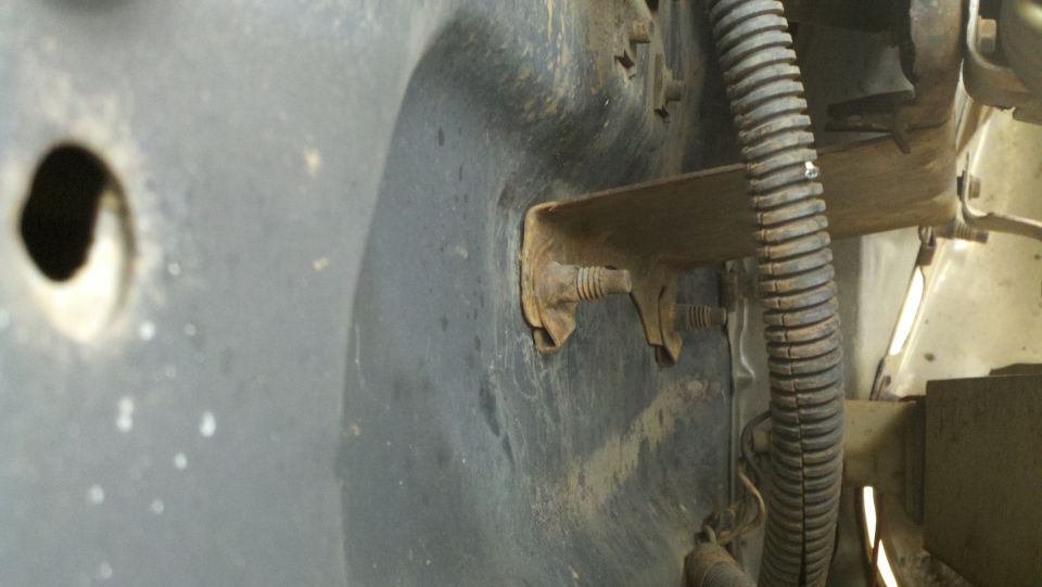 Close-up of a rusty metal bracket bolted to a dark, possibly plastic, fender liner with a hole in it. Corrugated wire loom is visible