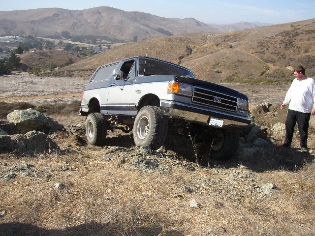 Lifted blue and silver Ford Bronco flexing over rocks on a dry hillside trail