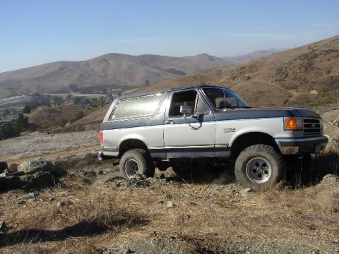 1989 Ford Bronco - A two-tone silver and blue Ford Bronco navigates a , dry terrain under a clear blue sky.