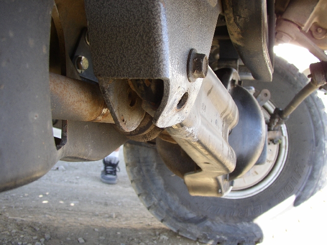 Underbody view showing rusty exhaust pipe, bracket, and tire over gravel