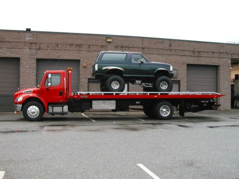 1994 Ford Bronco - A dark green Ford Bronco is transported on a red flatbed tow truck in front of a brick building.