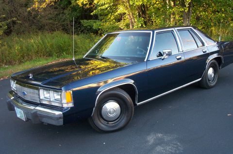 1991 Ford LTD Crown Victoria - A dark blue sedan with chrome accents sits parked on a paved surface, with foliage visible behind it.