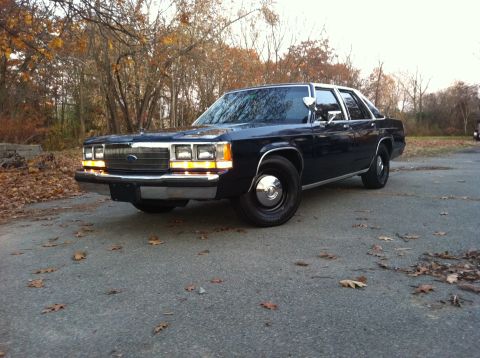 1991 Ford LTD Crown Victoria - A dark navy blue full-size Ford sedan photographed from a front three-quarter angle on a leaf-strewn asphalt lot, featuring a chrome grille…
