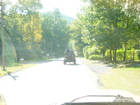 1979 Ford Bronco - A dark SUV with a roof rack travels down a tree-lined road on a bright, sunny day.