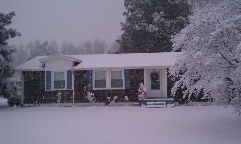 1979 Ford Bronco - A house sits covered in snow during a winter storm, with trees and bushes also blanketed.
