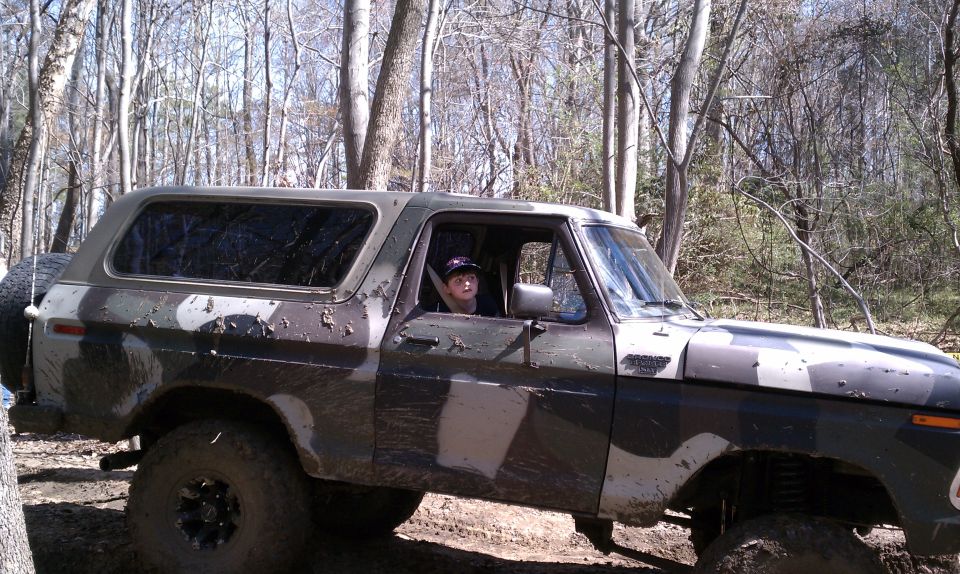 Camouflaged Ford Bronco with spare tire on rear, driven by a young person in a wooded area